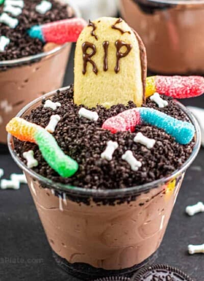 Close up of a chocolate dirt pudding cup decorated with gummy worms, bone sprinkles and a cookie headstone.