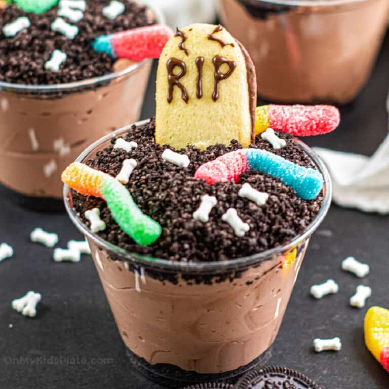 Close up of a chocolate dirt pudding cup decorated with gummy worms, bone sprinkles and a cookie headstone.