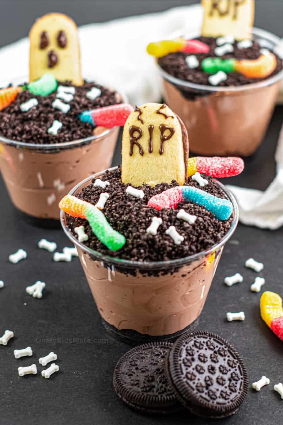 Three clear cups filled with pudding, topped with Oreo crumbs and tombstone and ghost shaped cookies.
