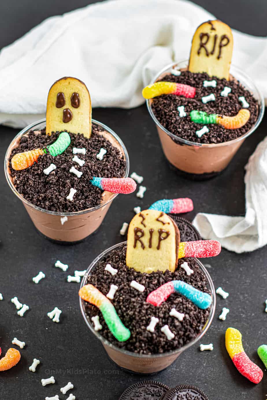 Three finished Graveyard dirt pudding cups topped with headstones, ghosts, gummy worms and bone candy sprinkles.
