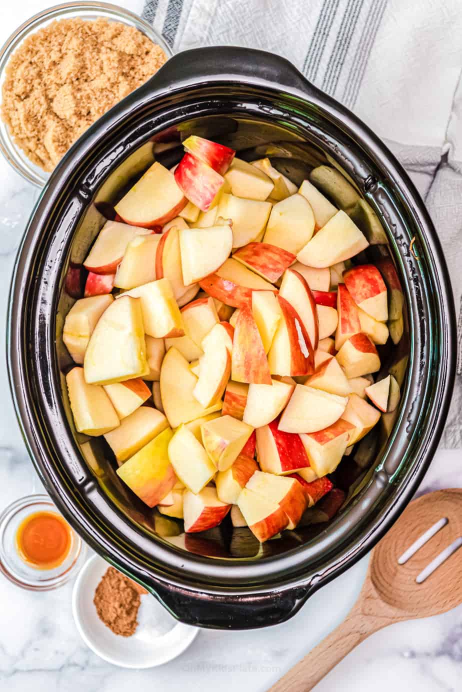 Sliced apples in a crockpot with other apple cutter ingredients in bowls nearby.
