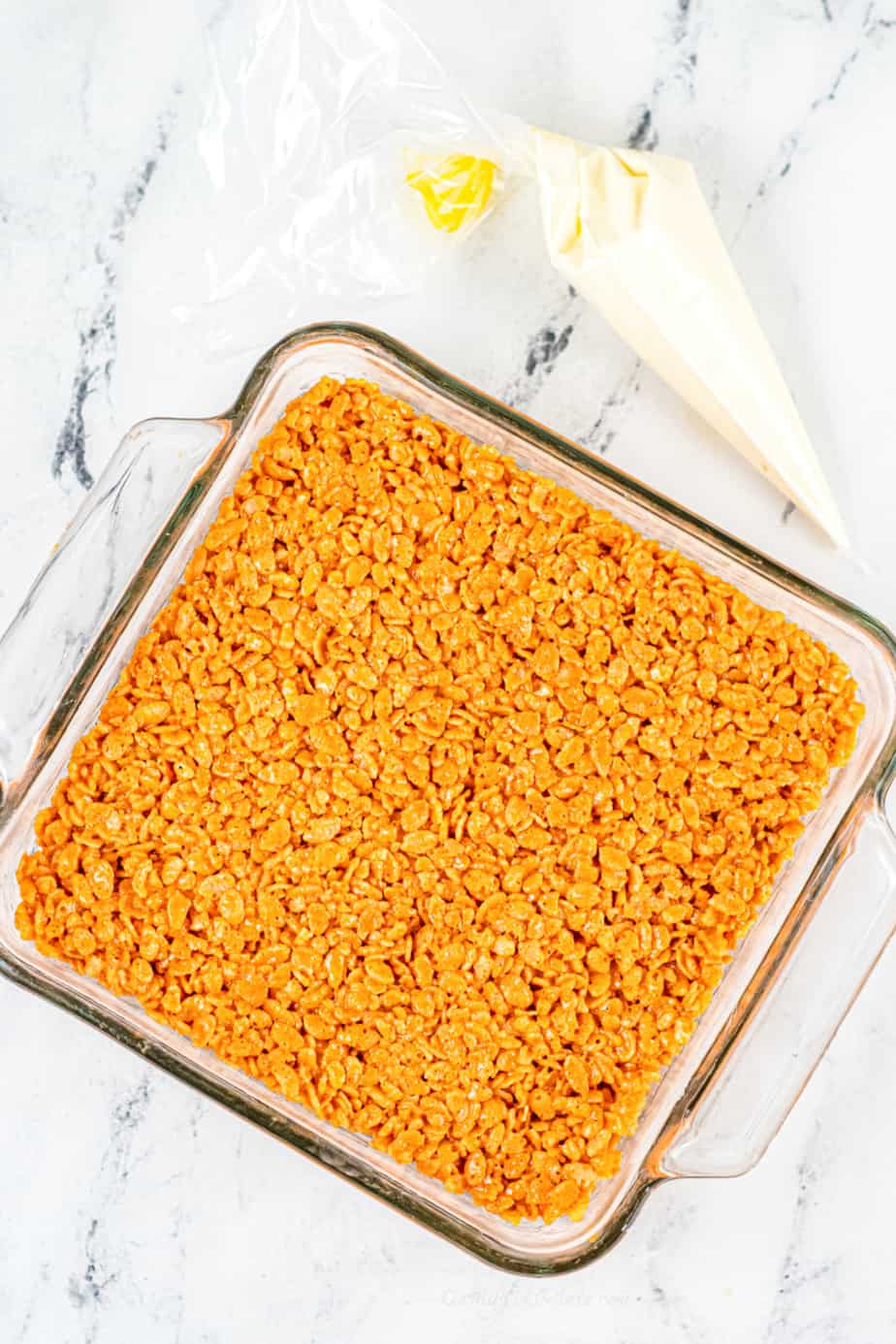 Pressing the pumpkin rice krispie mixture into a square glass pan with a pastry bag of melted chocolate next to the pan.