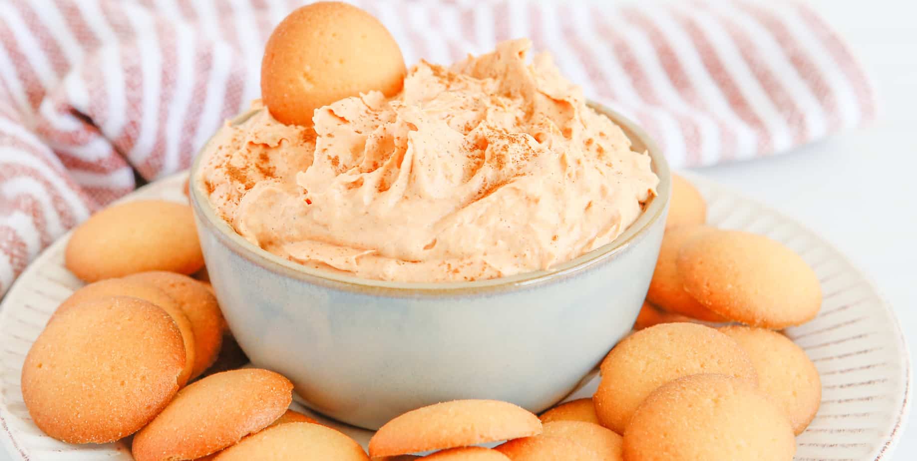 Wide side view of creamy pumpkin dip in a bowl with cookies on a plate surrounding the bowl.