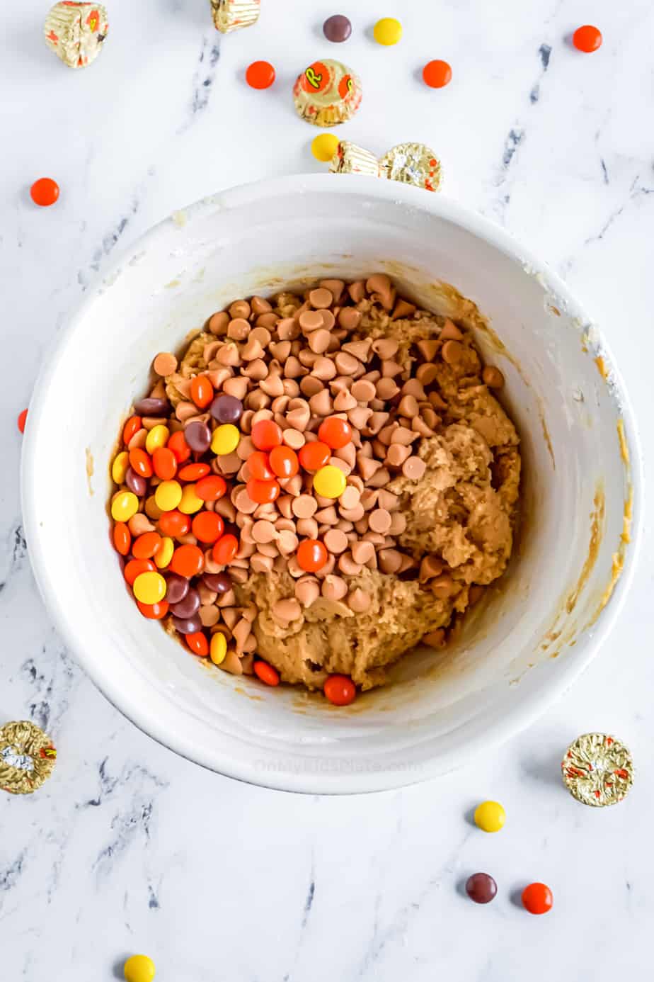 Cookie dough in a bowl from overhead with Reese's Pieces and peanut butter chips on top about to be mixed in.