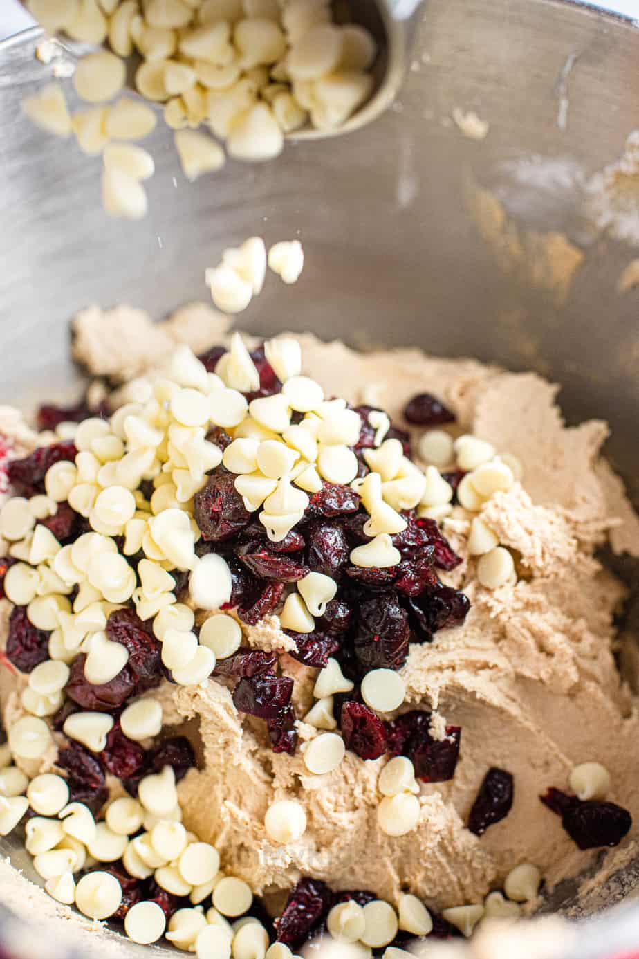 White chocolate chips and dried cranberries being poured close up into a bowl full of cookie dough.