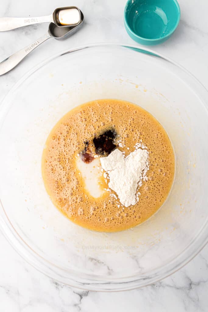 A glass bowl with mini pecan pies filling being mixed with flour, vanilla extract, and milk on a marble surface, with measuring spoons and a small blue bowl nearby.