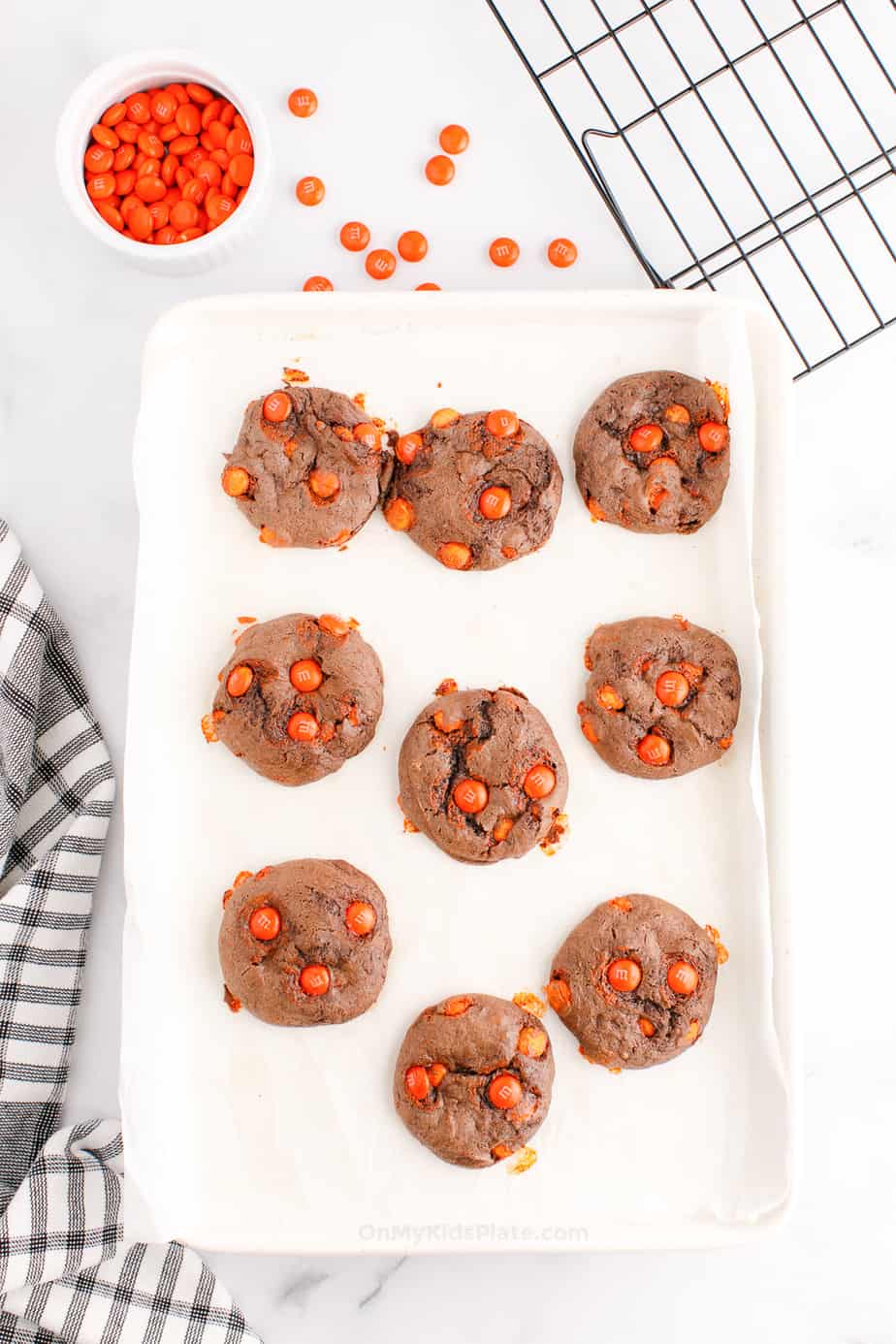 Chocolate cookies with orange candies baked on a pan with a wire rack and more orange candies in a bowl next to the pan