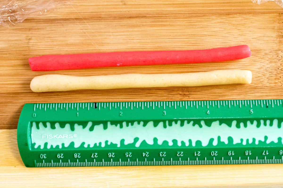 Red and white cookie dough rolled into ropes being measured by a ruler on a cutting board.