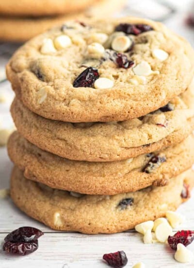 Close side view of four cranberry white chocolate cookies stacked with more cookies and extra chocolate and cranberries scattered nearby.