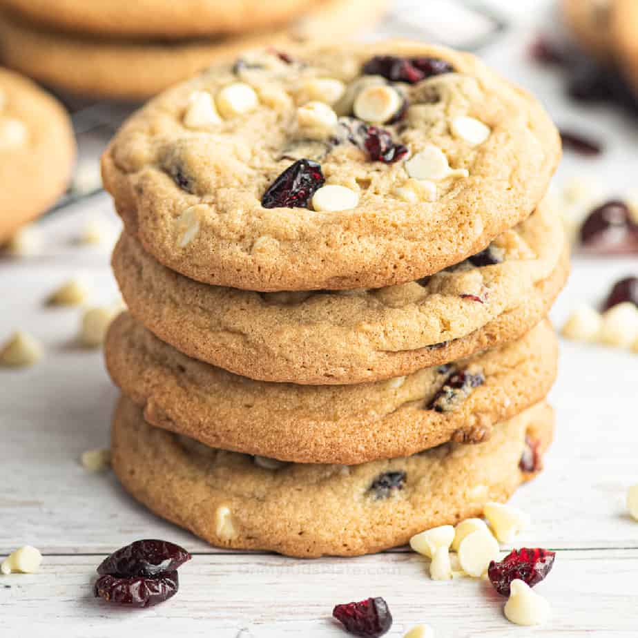 Close side view of four cranberry white chocolate cookies stacked with more cookies and extra chocolate and cranberries scattered nearby.