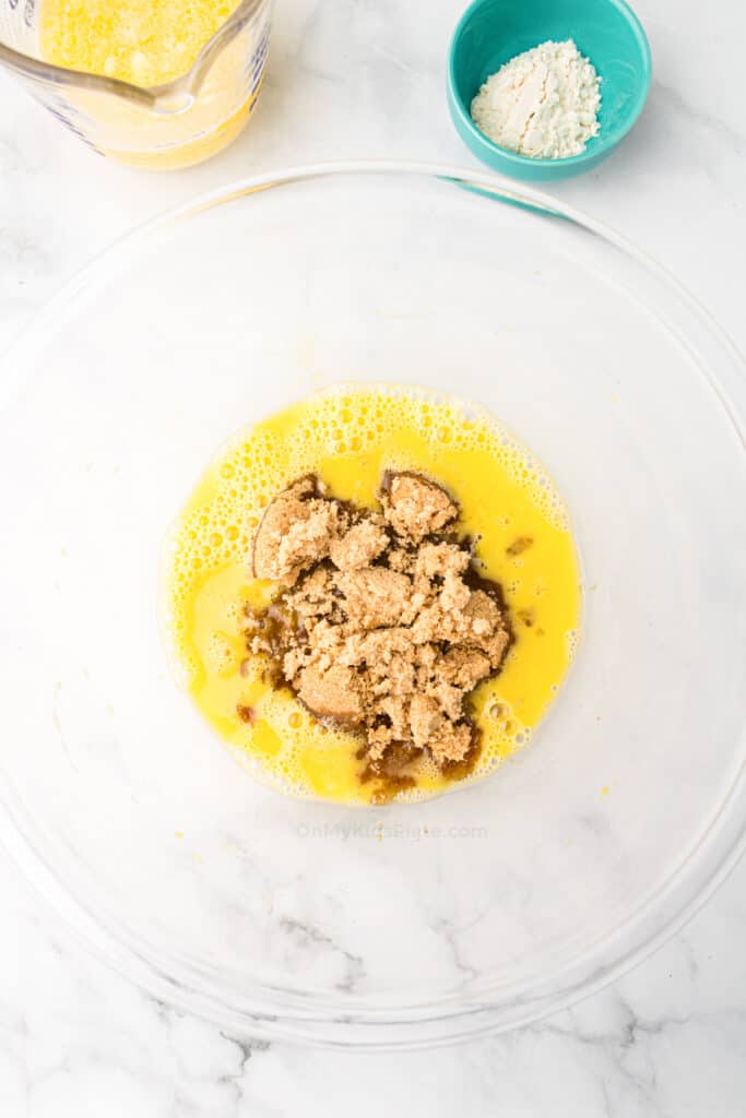 A clear glass bowl contains whisked eggs and brown sugar for mini pecan pie. Nearby are a small bowl of flour and a measuring cup with more whisked eggs on a white marble surface.