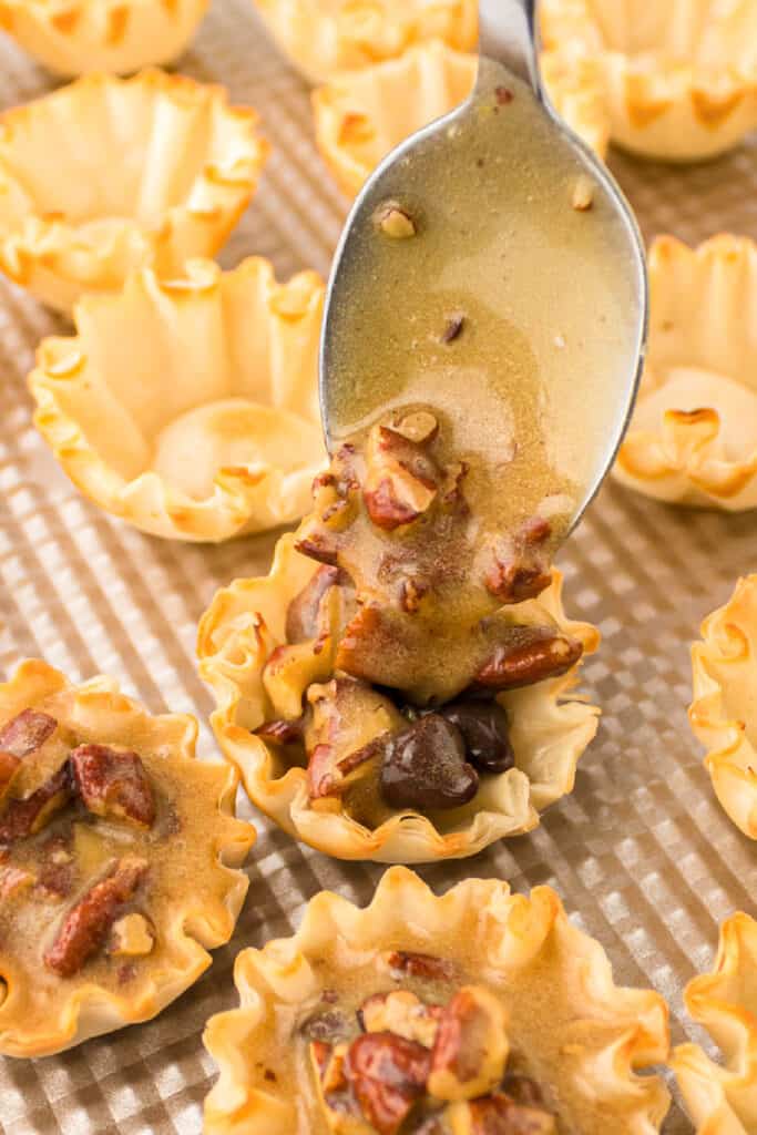 A spoon fills mini phyllo pastry shells with a rich mini pecan pie and syrup mixture, surrounded by other empty and partially filled shells on a baking sheet.