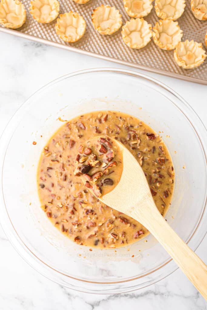 A glass bowl with mini pecan pie filling being mixed with a wooden spoon; empty tart shells are visible on a baking tray in the background.