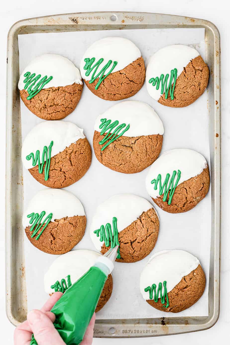 Molasses cookies dipped in white chocolate on a pan with green squiggle chocolate Christmas trees being added with a piping bag.