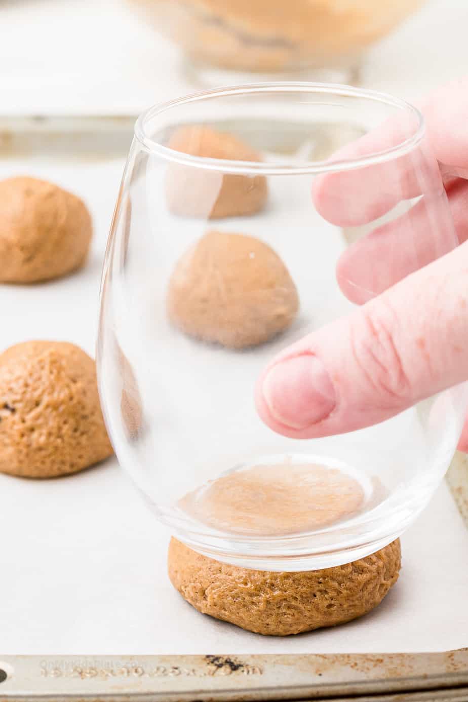 Side view of a glass pressing down on molasses cookie dough ball on a cookie sheet.