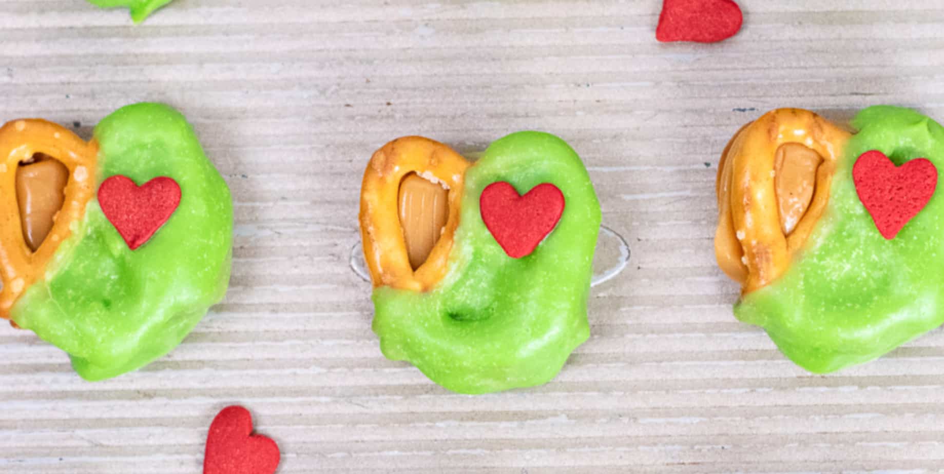 Three pretzels with caramel on a pan decorated with green candy melts and a red heart sprinkle