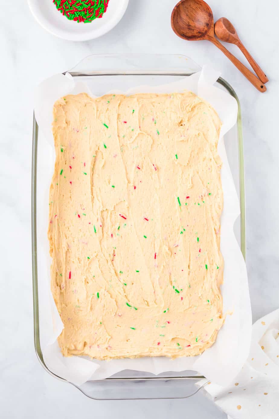 Sugar cookie dough full of red and green sprinkles pressed into a parchment lined glass rectangular pan from overhead.