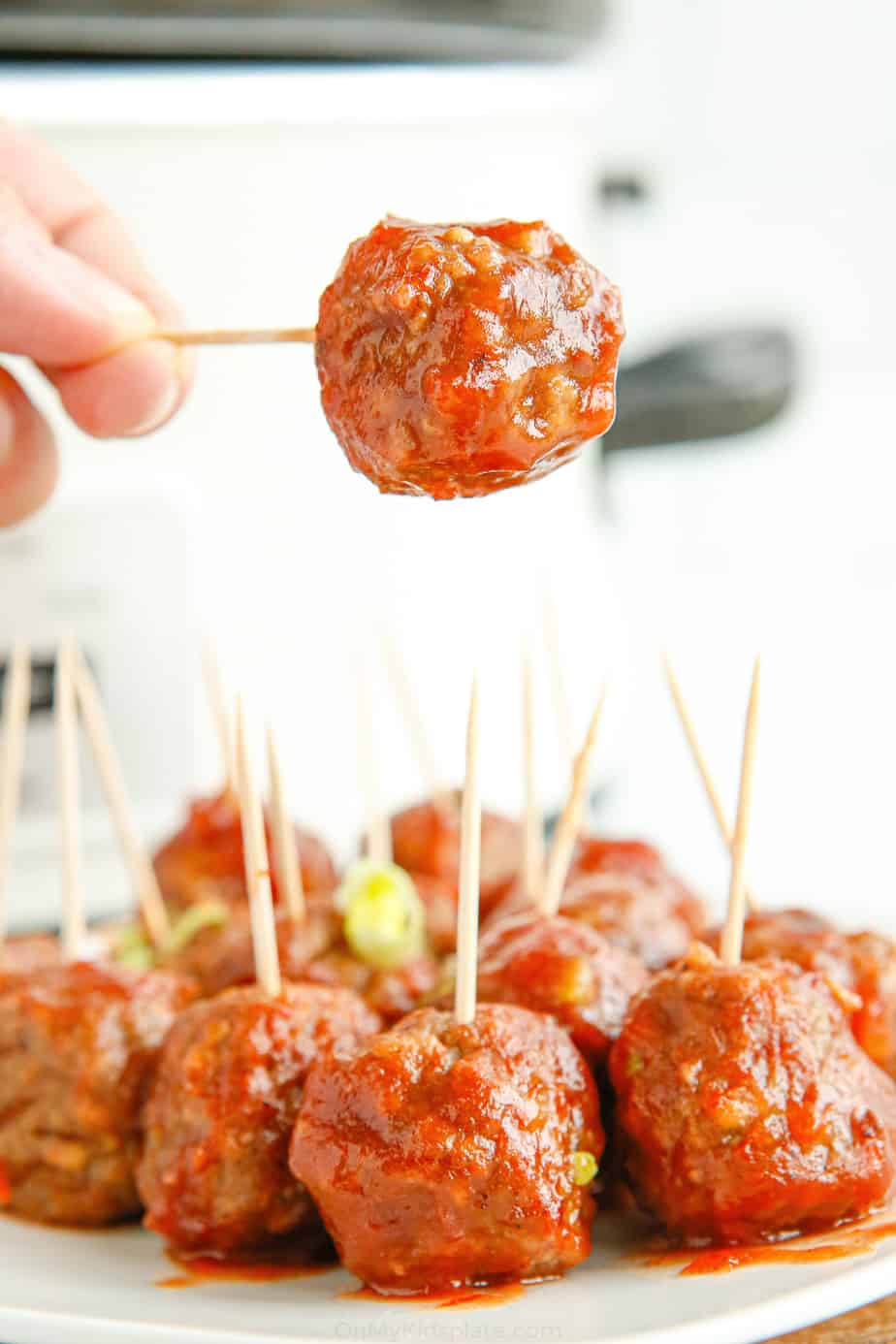 Meatballs in sauce on a plate with a toothpick in each meatball. One meatball on a toothpick is being taken from the platter.