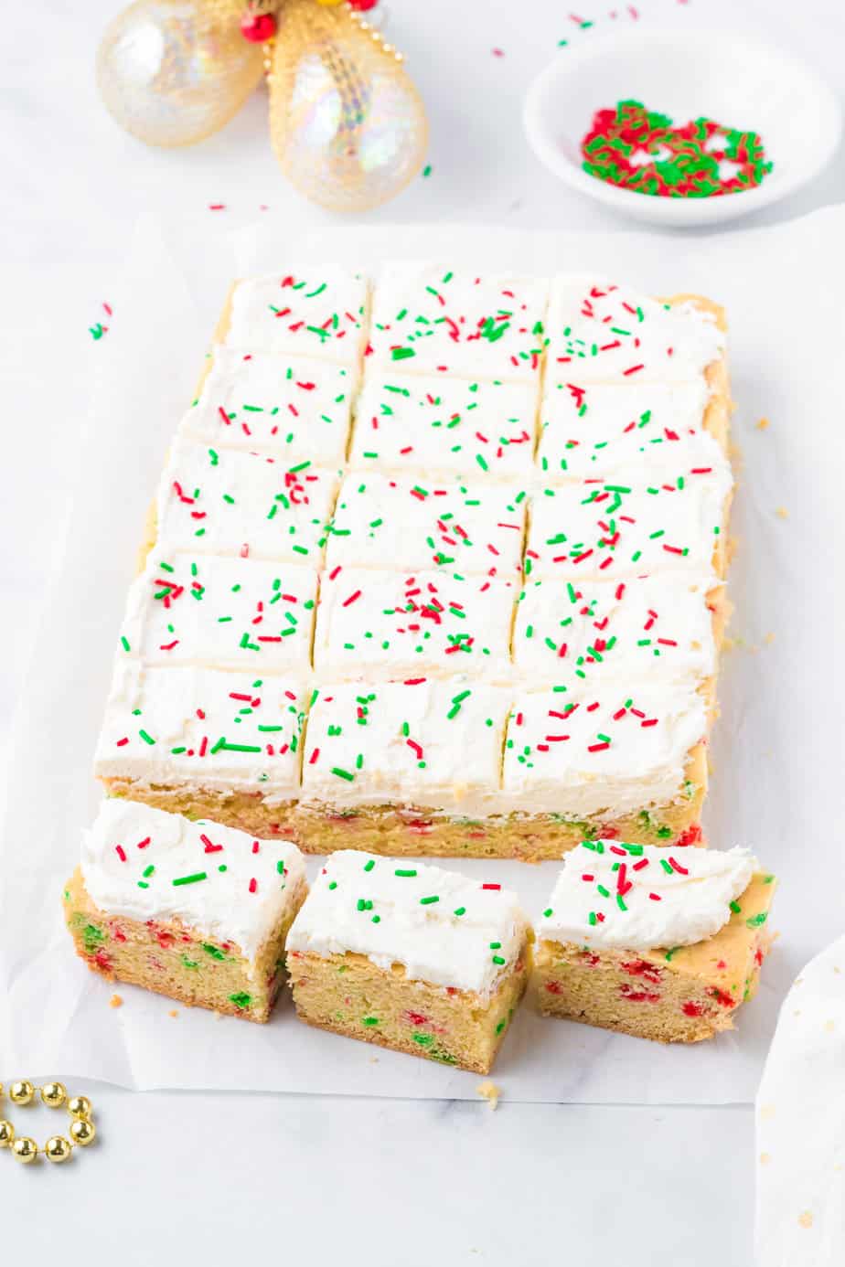 Frosted and sliced sugar cookie bars on parchment from the side with red and green sprinkles on top and in a nearby bowl.