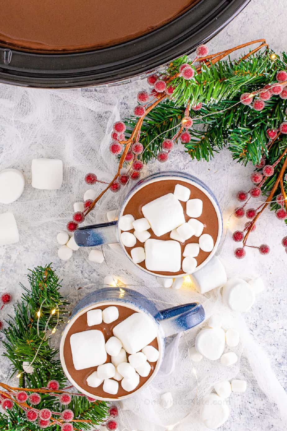 Overhead two mugs of hot chocolate with marshmallows next to the slow cooker near holiday garlands