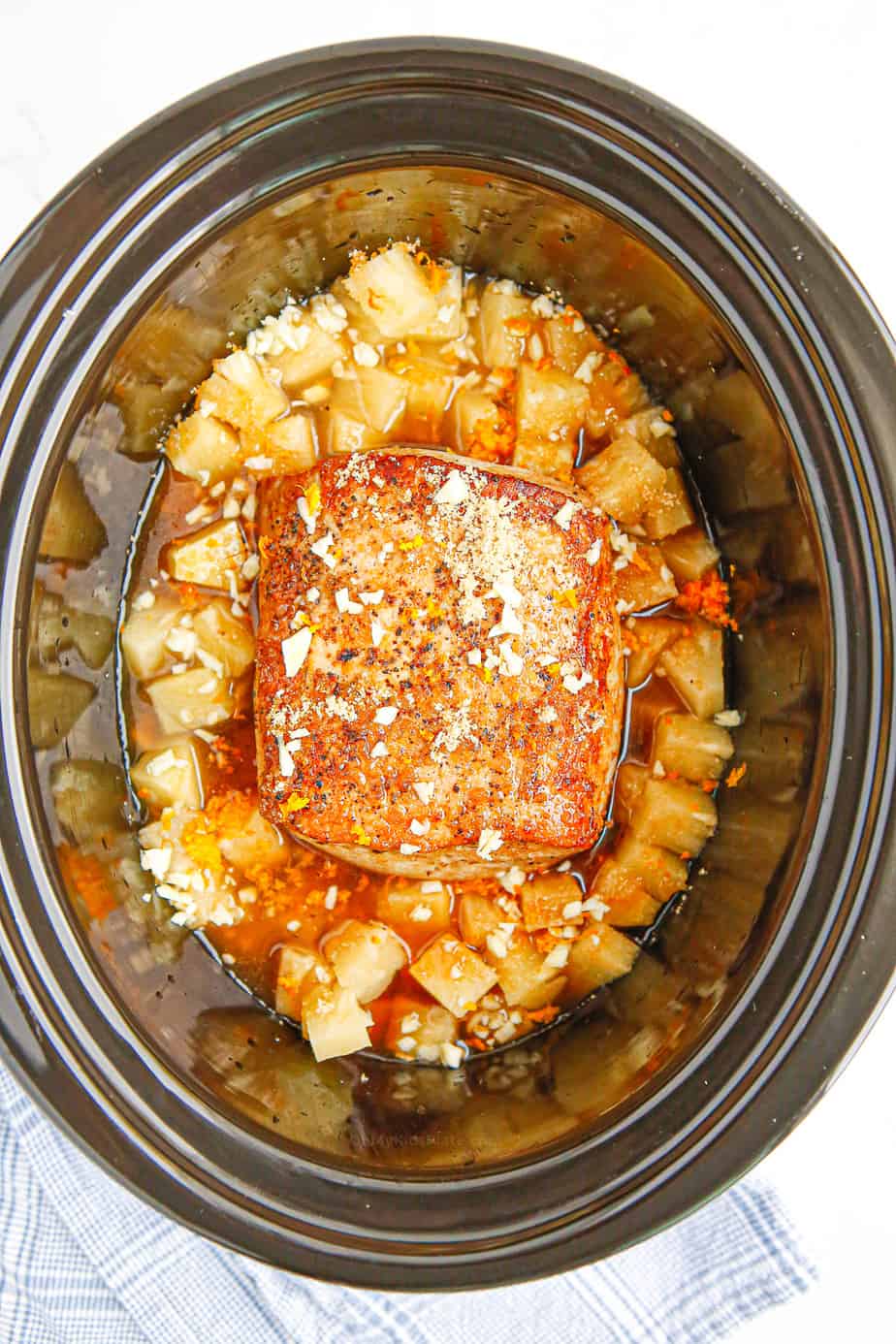 Browned pork loin in a slow cooker from above surrounded by pineapple and spices