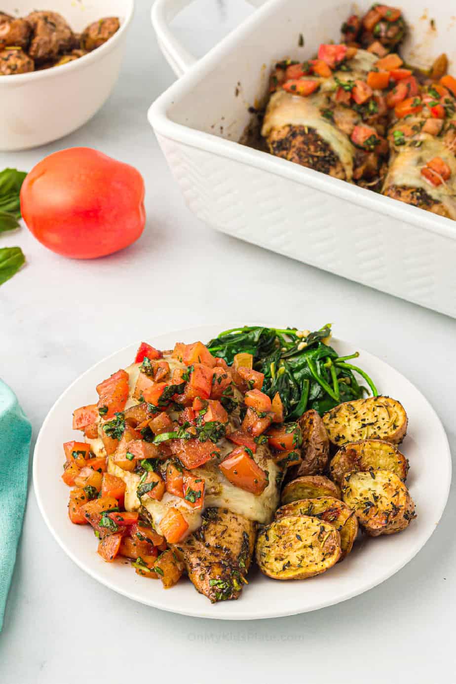 Baked bruschetta chicken on a plate on a table with vegetables and a pan of more chicken behind
