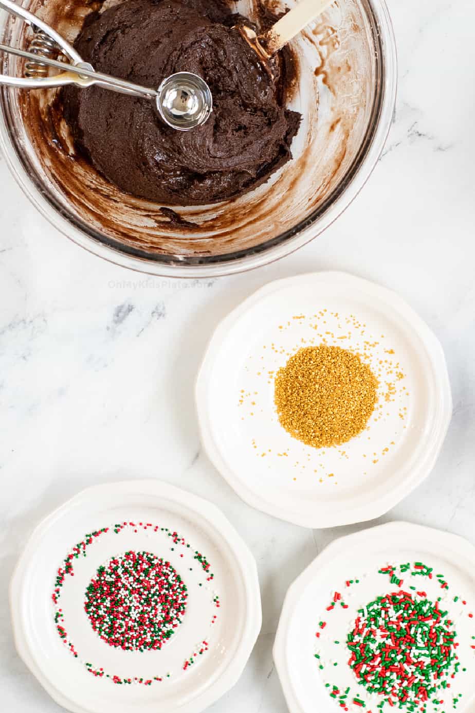 Bowl of brownie truffle dough with a cookie scoop and three plates full of sprinkles