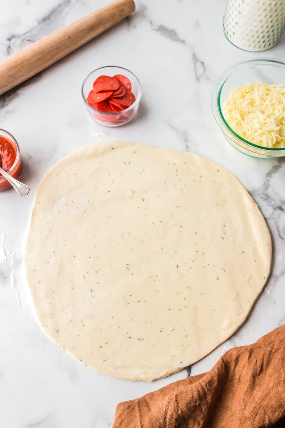 pizza dough rolled flat on a counter with other ingredients in bowls nearby