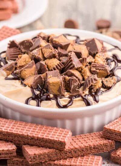 Square image of cheesecake dip close up topped with peanut butter cups and chocolate in a bowl next to chocolate cookies