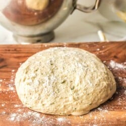 Pizza dough on a cutting board with flour and a mixer bowl behind