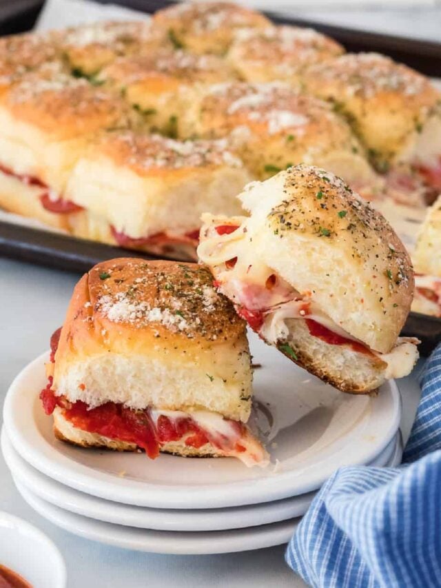 Two pizza sliders stacked on a plate from the side with a pan full of sliders behind