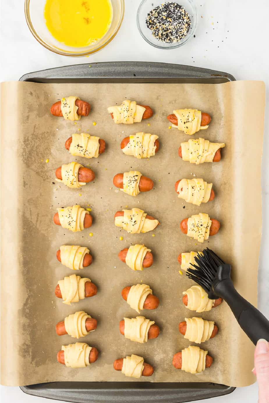 brushing pigs in a blanket on a pan with egg wash and adding seasoning