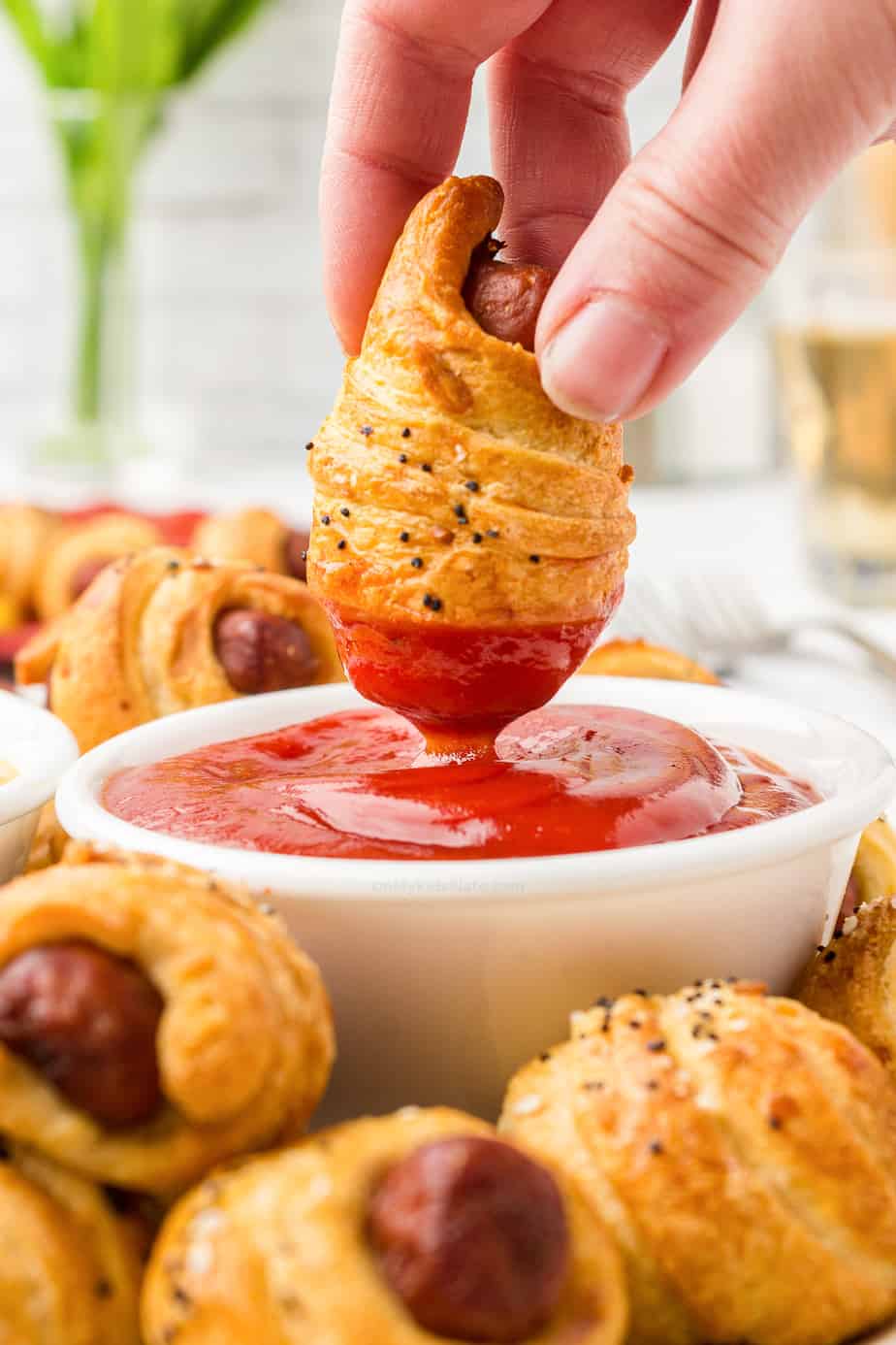 Dough wrapped sausage being dipped into ketchup in a bowl on a platter of pigs in a blanket