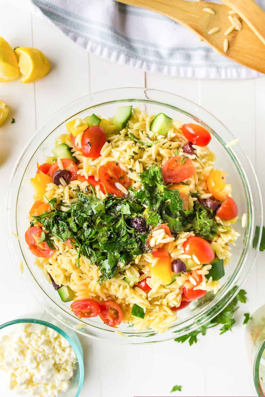 Adding fresh herbs and dressing to a bowl of orzo mixed with vegetables from overhead