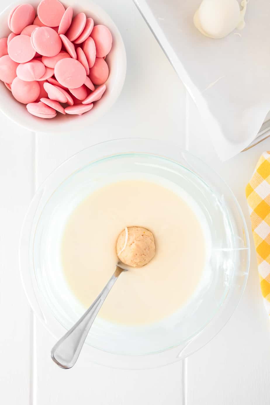 Dipping strawberry lemonade truffles in a bowl of melted white chocolate with a fork. A pan of more truffles and a bowl of pink candy melts sit nearby
