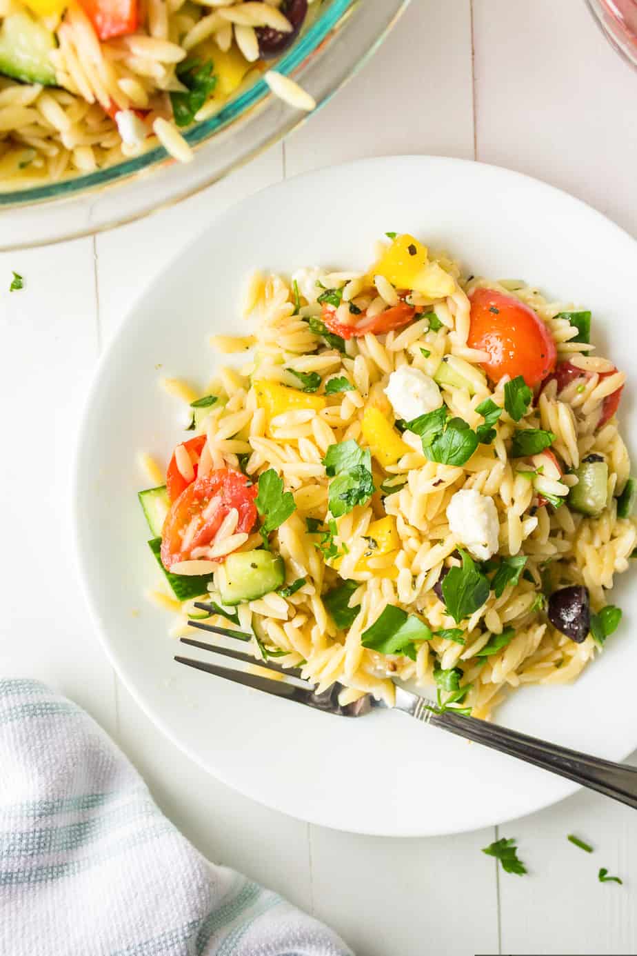 Greek orzo salad on a plate from overhead with a fork next to a bowl of more cold salad topped with feta cheese, parsley, and mixed with colorful diced vegetables