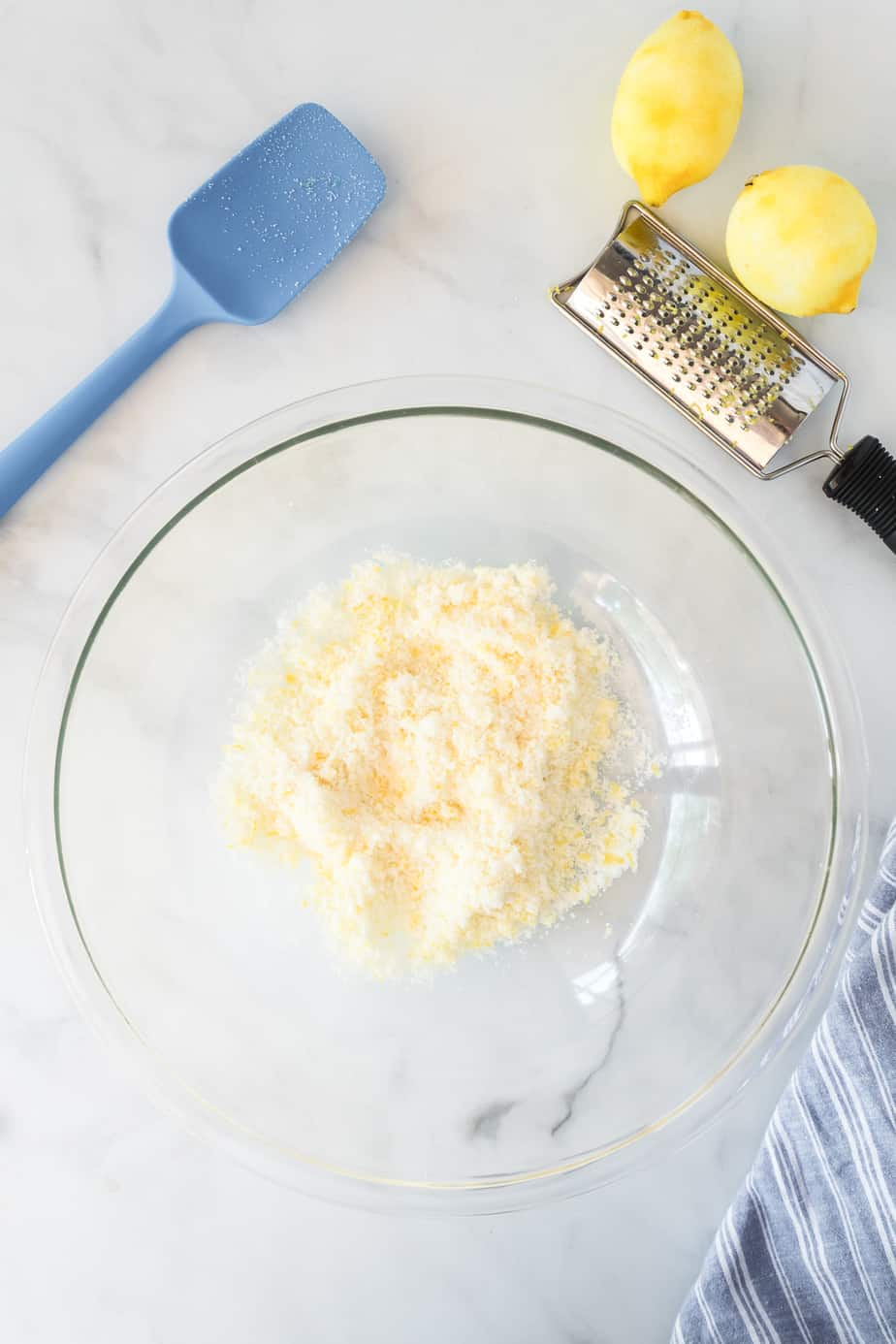 Lemon zest being mixed into white sugar in a large bowl from overhead with lemons and a zester and a silicone spatula nearby on the counter
