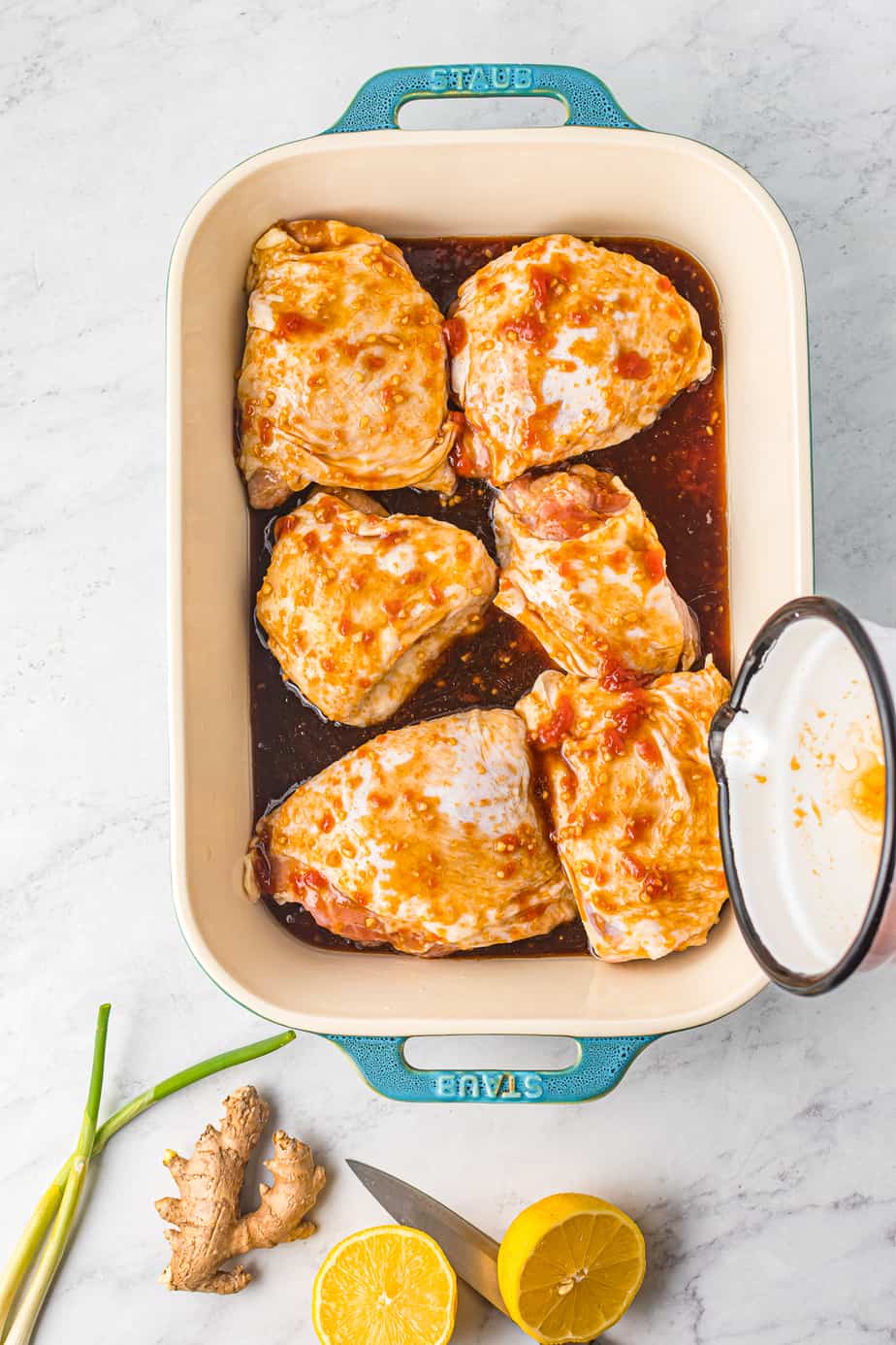 A cup pours honey garlic marinade over bone in chicken thighs in a baking dish from overhead
