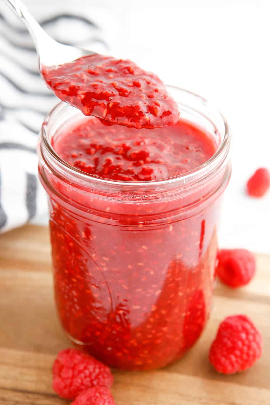 Raspberry jam being lifted from a jar with a spoon from the side. Fresh raspberries are scattered on the wood cutting board beneath the jar
