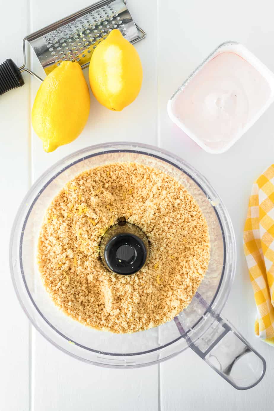 vanilla sandwich cookie crumbs in a food processor from overhead with lemons, a lemon zester and strawberry cream cheese nearby