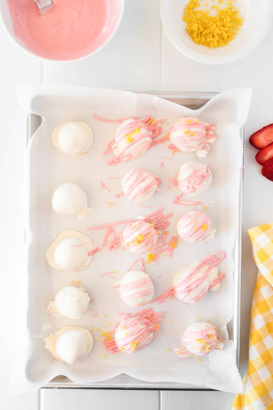 strawberry lemonade truffles dipped in white chocolate on a pan being drizzled with melted pink chocolate and lemon zest from nearby bowls.