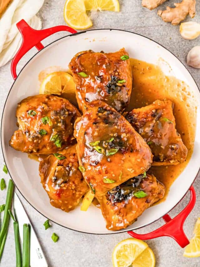 Baked honey garlic chicken thighs in sauce in a serving platter with leftover green onion, ginger and lemon slices nearby