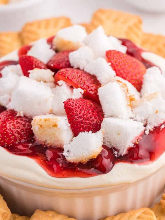 Close up side view of cheesecake dip topped with fresh strawberries, strawberry pie filling and angel food cake in a serving bowl surrounded by cookies