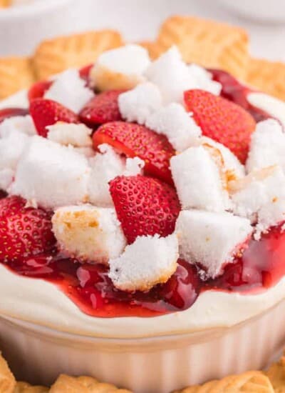 Close up side view of cheesecake dip topped with fresh strawberries, strawberry pie filling and angel food cake in a serving bowl surrounded by cookies