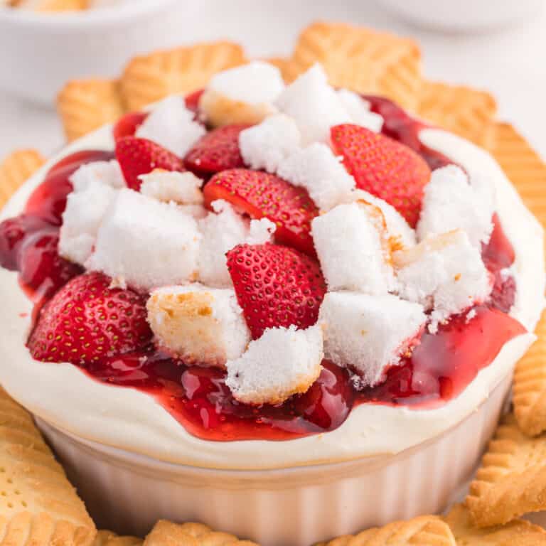 Close up side view of cheesecake dip topped with fresh strawberries, strawberry pie filling and angel food cake in a serving bowl surrounded by cookies