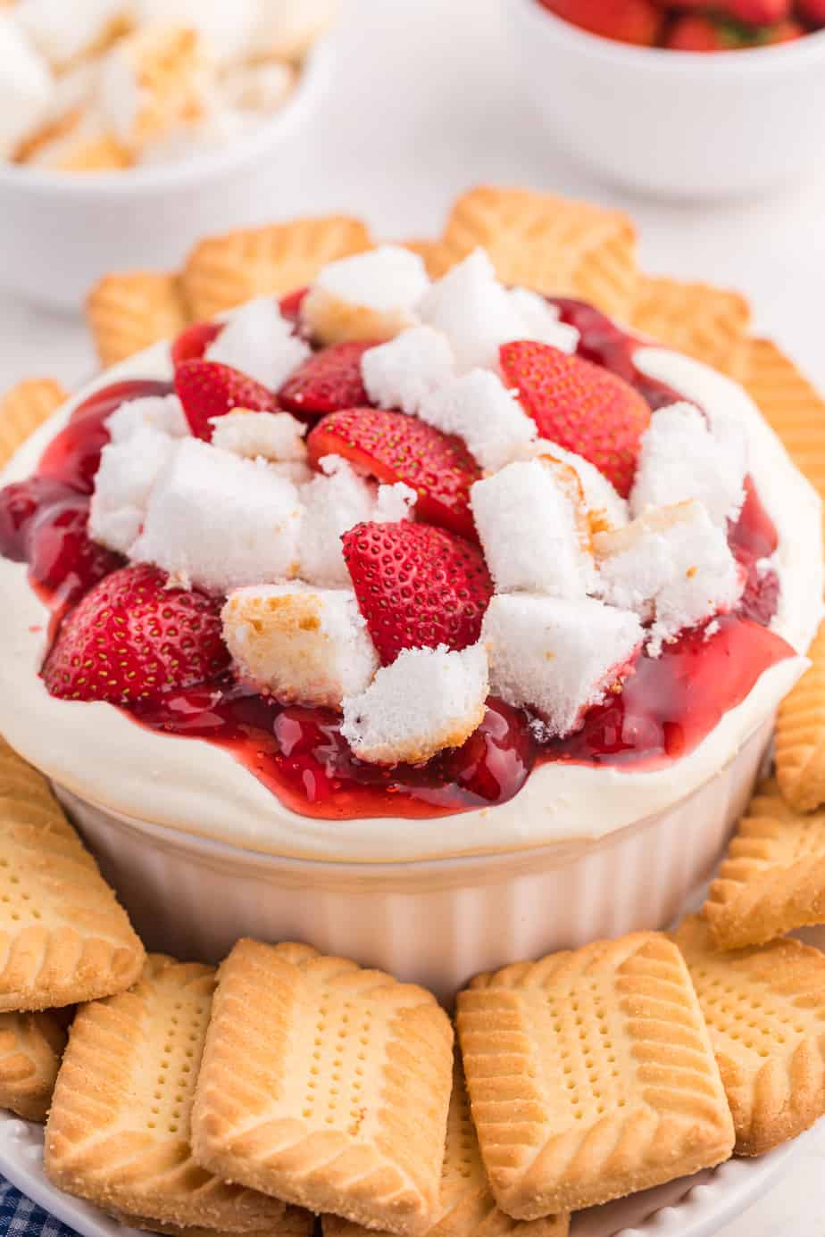 Cheesecake dip topped with strawberries, red strawberry sauce and angel food cake pieces surrounded by cookies on a platter