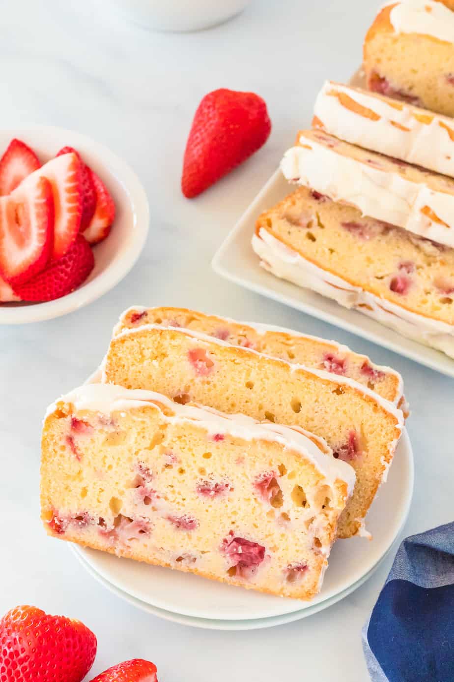 Two slices of strawberry bread with glaze on a plate next to a platter of quick bread and a bowl of fresh strawberries