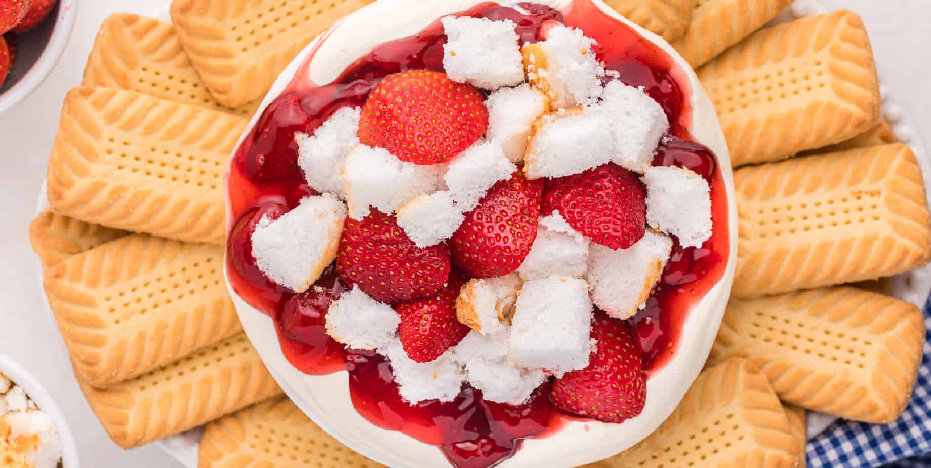 wide view of cheesecake dip in a bowl topped with strawberry sauce, fresh strawberries, pieces of angel food cake all on a platter with vanilla cookies