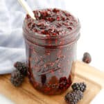 blackberry jam from the side in a jar that is open with a spoon in the top and blackberries scattered around the bottom of the jar
