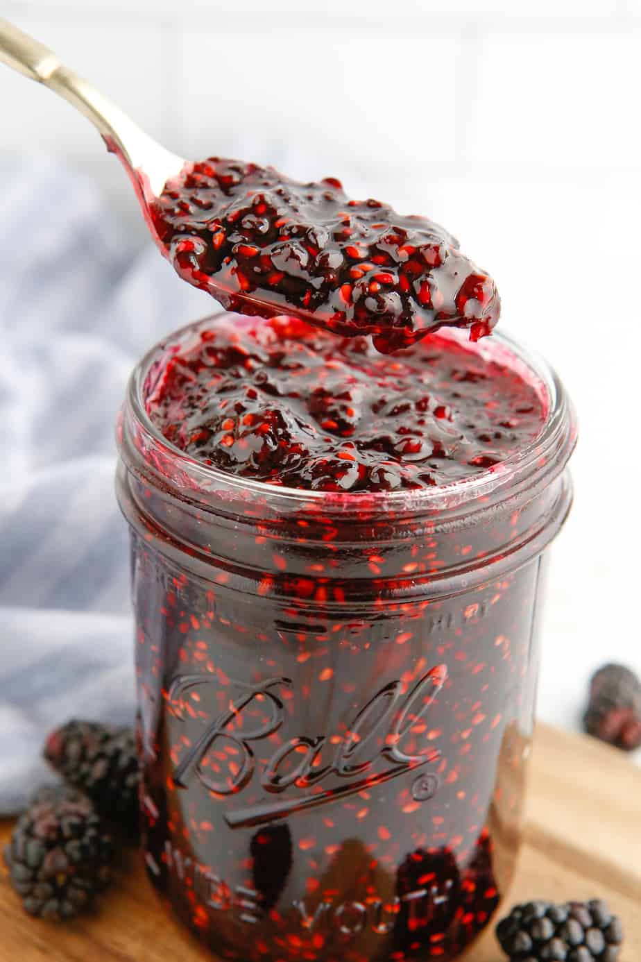A spoon pulling a spoonful of blackberry jam from the top of a jar of jam from the side with fresh blackberries around the bottom of the jar.